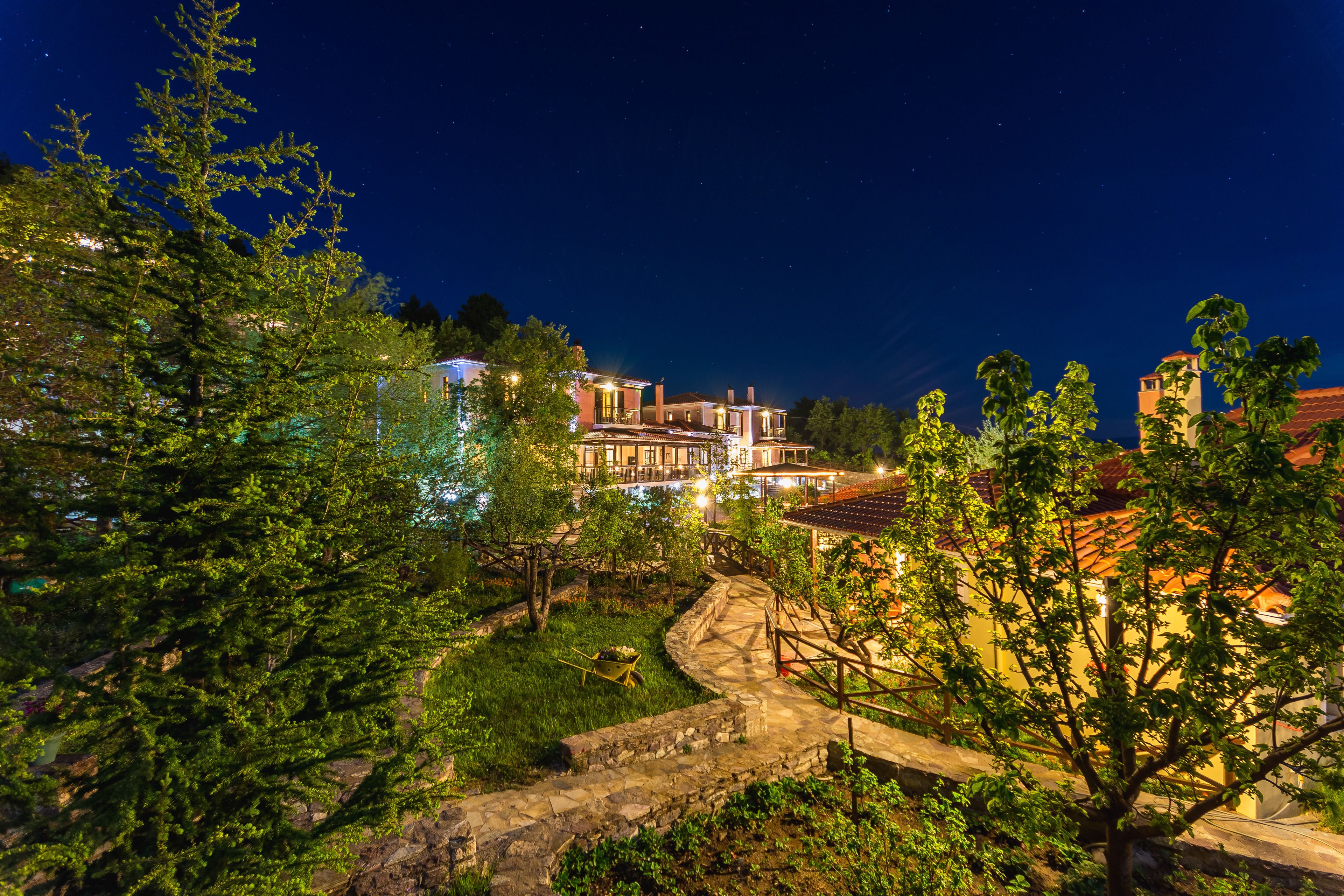 A well-lit, multi-story building with a stone pathway leading to a pool area, surrounded by lush greenery.