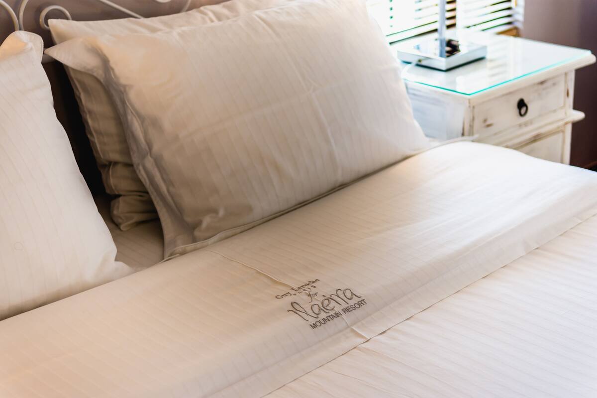 Ein ordentlich bezogenes Bett mit weißer Decke und Kissen, ein Nachttisch mit Lampe sowie ein Schild mit dem Hotelnamen und der Adresse.