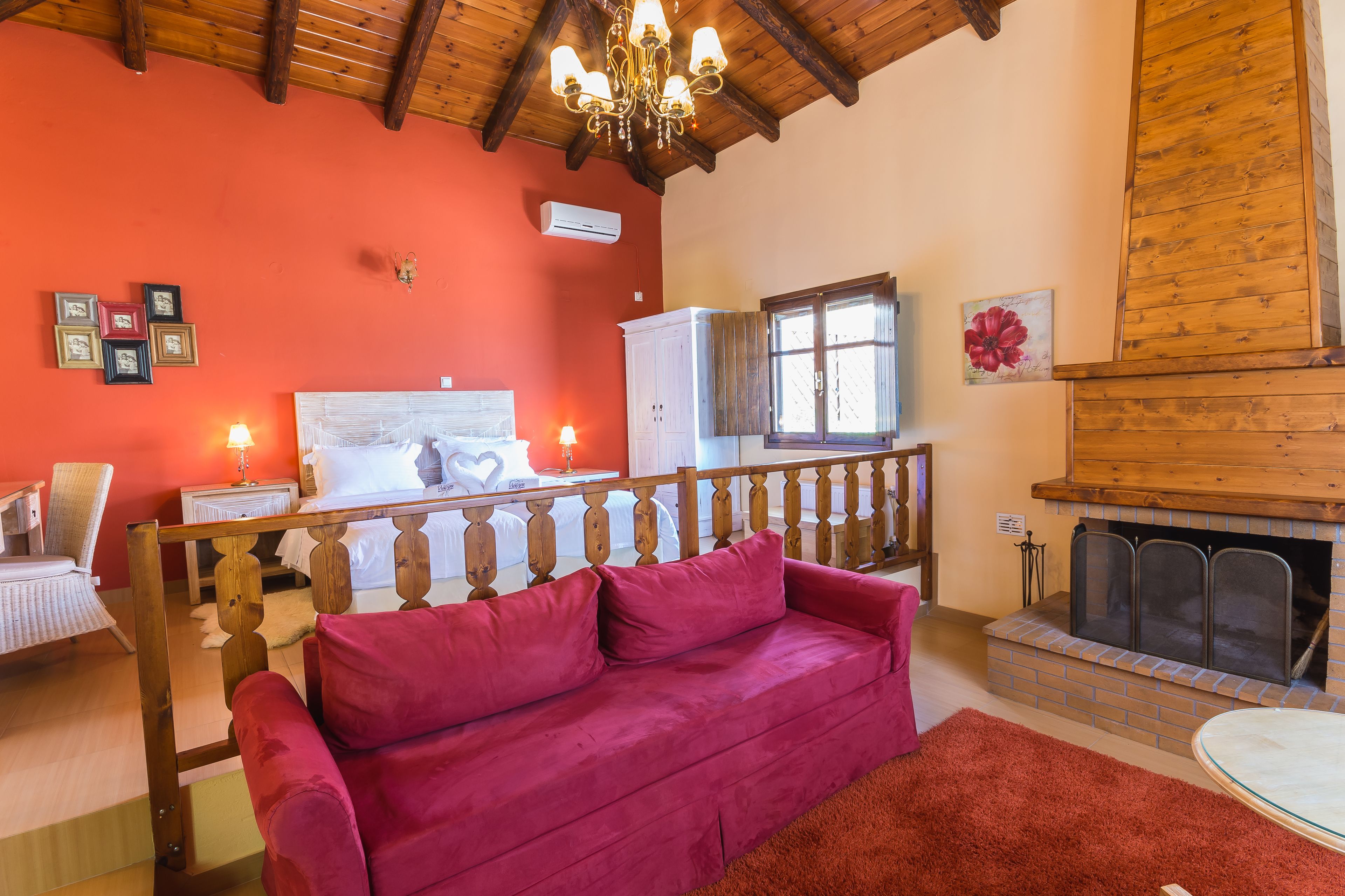 Una habitación acogedora con techo de madera, pared roja, cama con ropa blanca, sofá rosa y chimenea.