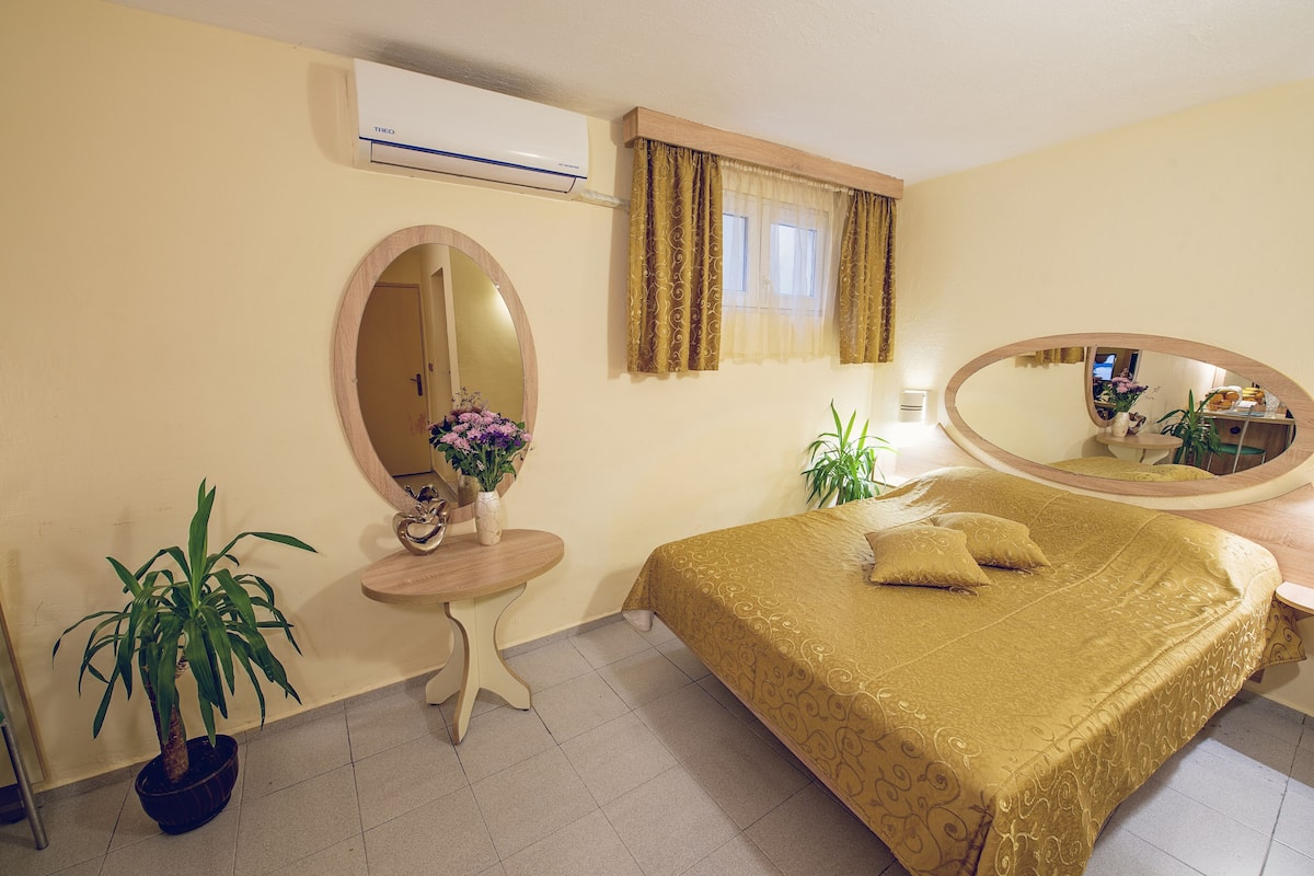 A hotel room with a bed, a round mirror, a small table with a vase of flowers, and a potted plant.