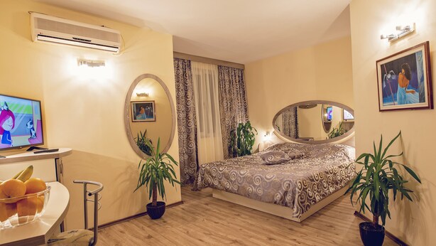 A bedroom with a bed, two potted plants, a TV, a desk with a bowl of fruit, and a mirror. - Hotel Color (Varna)