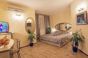 A bedroom with a bed, two potted plants, a TV, a desk with a bowl of fruit, and a mirror. - Hotel Color (Varna)