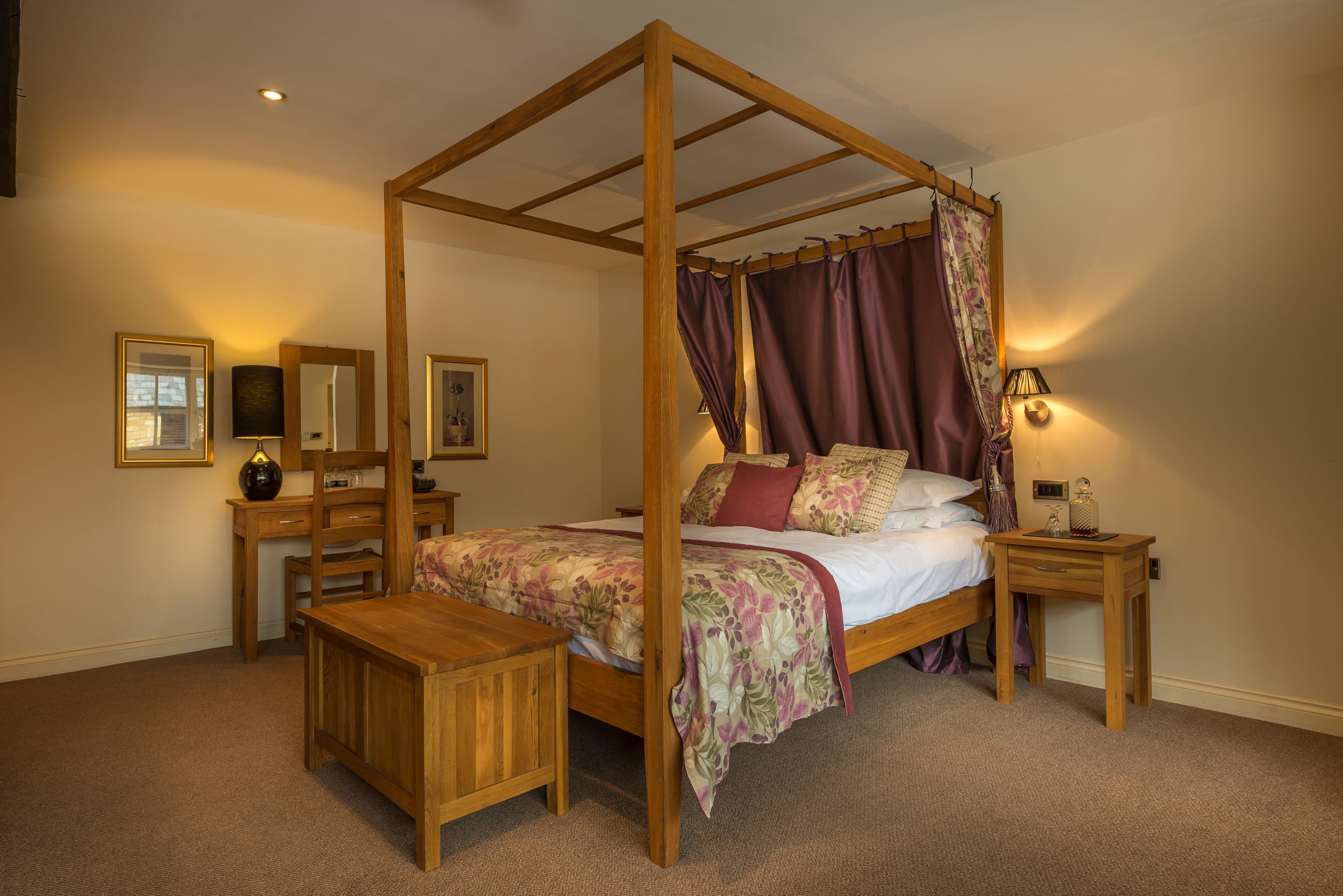 Ein Himmelbett mit floralem Überwurf, ein Nachttisch, ein Beistelltisch und ein Spiegel an der Wand.