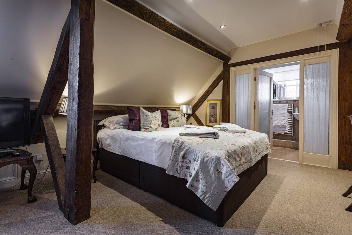 A bedroom with a large bed, wooden beams, a TV, and a bathroom visible through an open door.