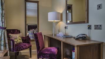 A hotel room with a wooden desk, two purple velvet chairs, a mirror, a lamp, a telephone, and a small cabinet.