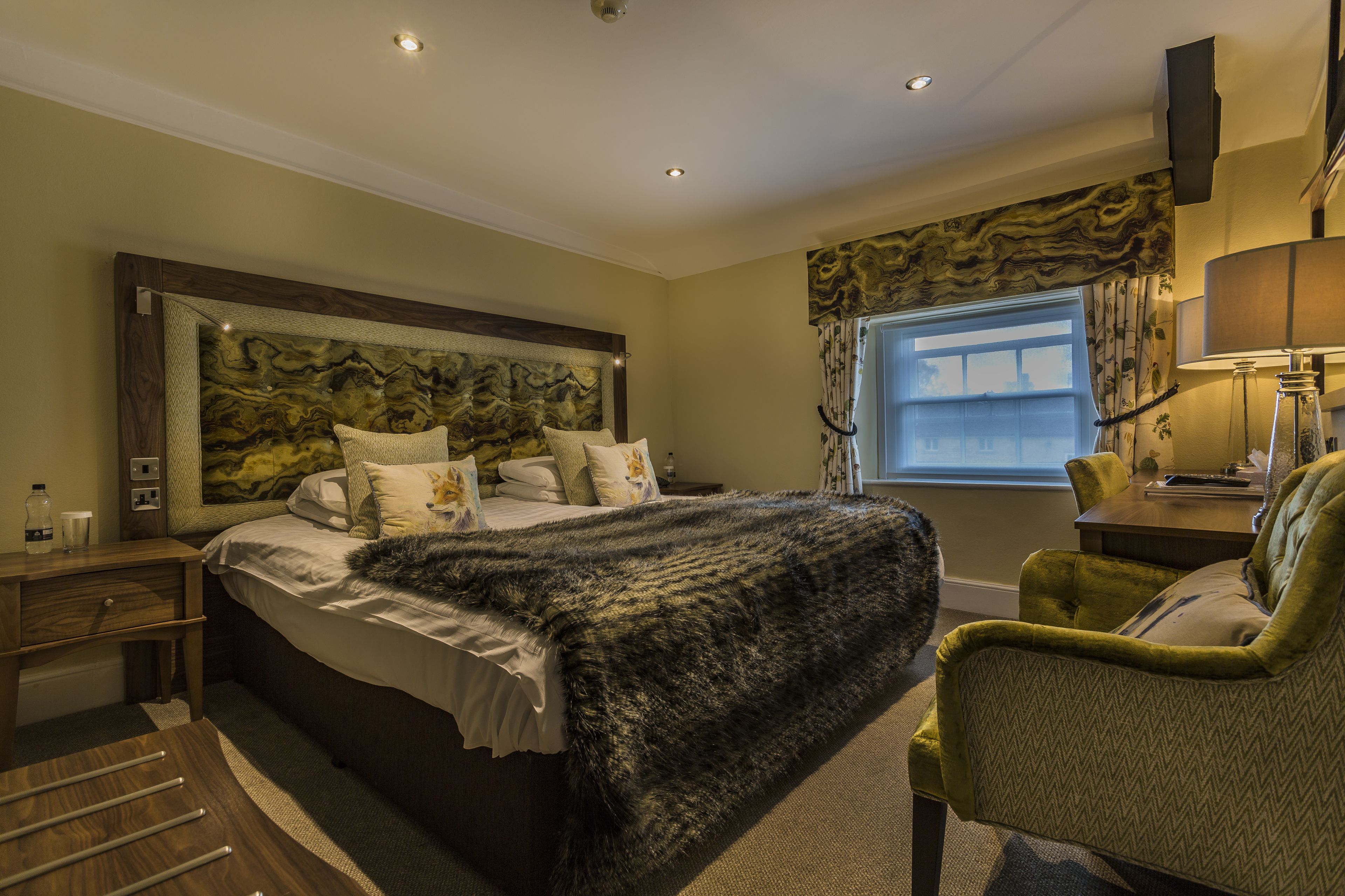 Een slaapkamer met een groot bed, een hoofdbord met een structuur, een nachtkastje met een lamp en een raam met gordijnen.