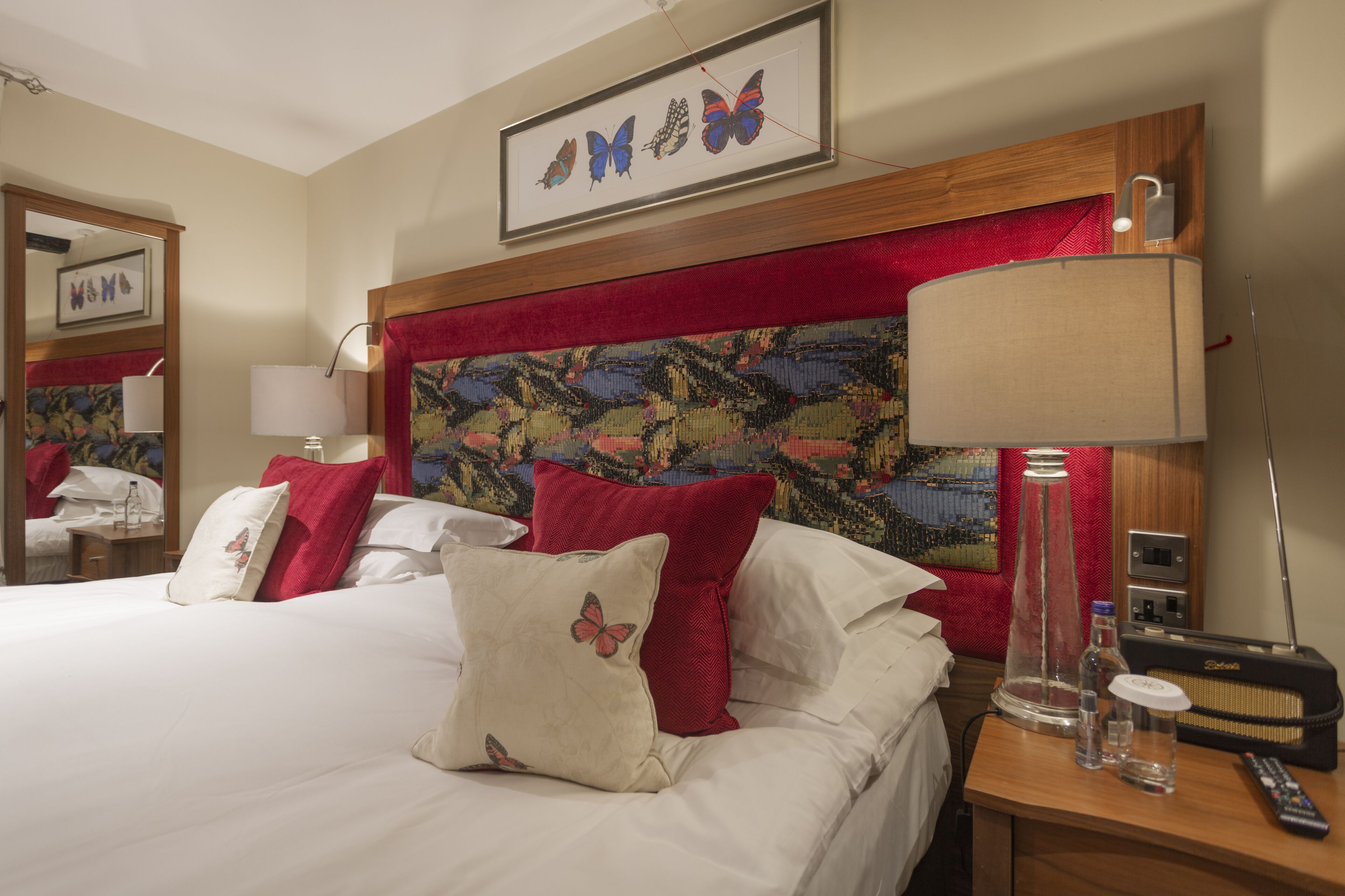 A hotel room with a large bed, red and white pillows, a painting on the headboard, a lamp, a radio, and a mirror.