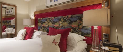 A hotel room with a large bed, red and white pillows, a painting on the headboard, a lamp, a radio, and a mirror.
