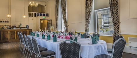 A dining area with multiple tables set for a formal event, featuring chandeliers and patterned curtains.