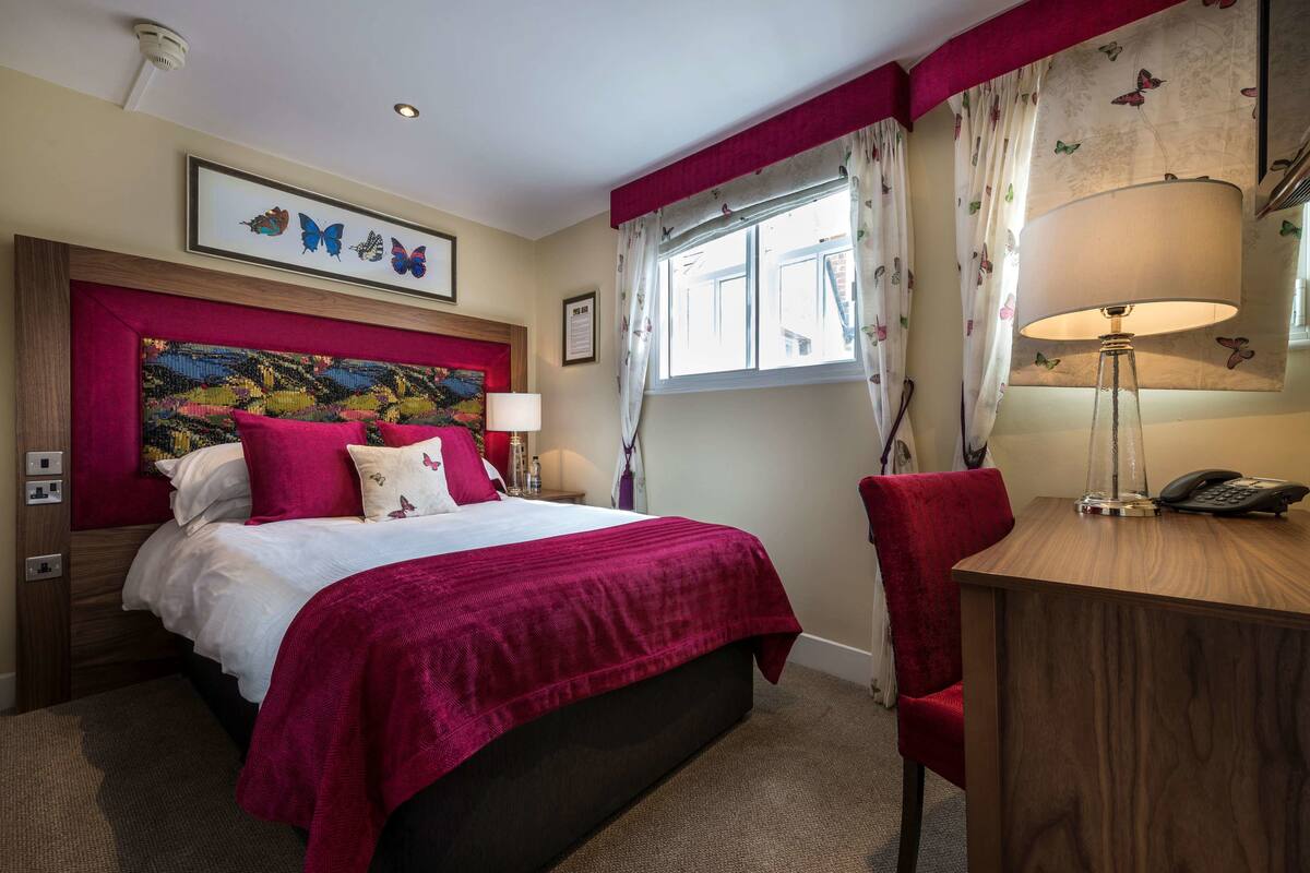 A hotel room with a bed, a desk, a chair, a lamp, and a window with curtains.