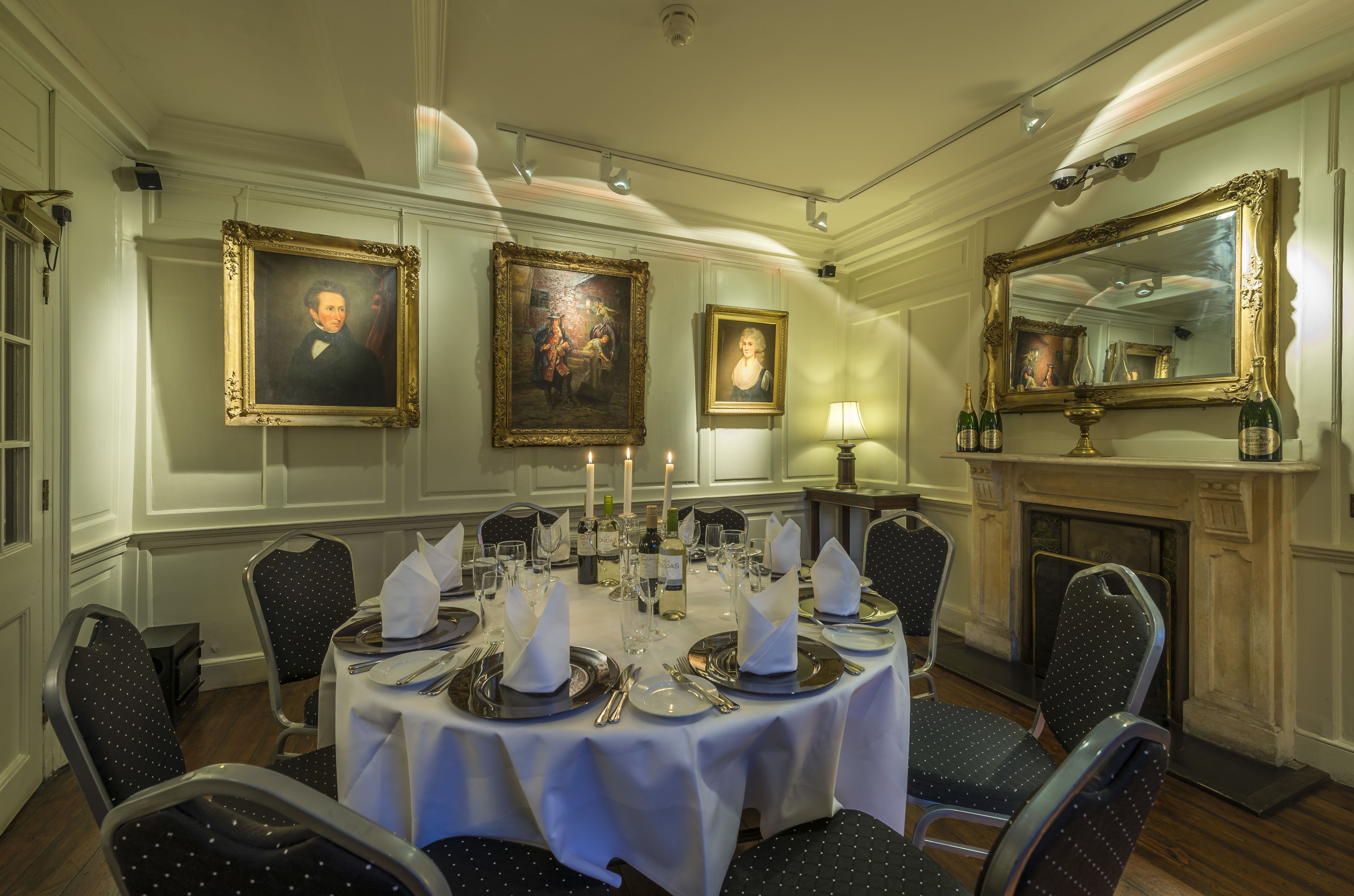 A formal dining room with a round table set for six, surrounded by chairs. There are framed portraits on the walls, a fireplace, and a mirror above the table.