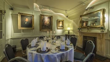 Un comedor formal con una mesa redonda dispuesta para seis personas, rodeada de sillas. Hay retratos enmarcados en las paredes, una chimenea y un espejo sobre la mesa.