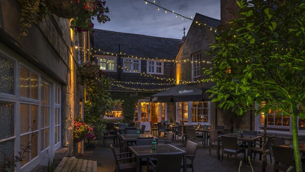 An outdoor seating area with tables and chairs, string lights, and a brick building with windows. - The White Hart Royal Hotel, Moreton-in-Marsh (Moreton-in-Marsh)