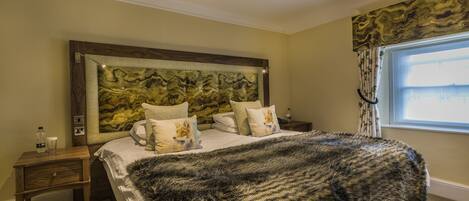 A bedroom with a large bed, a headboard with a decorative panel, a nightstand with a bottle and glass, and a window with curtains.