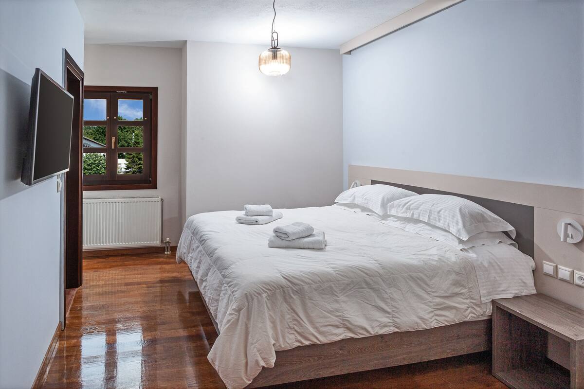 A bedroom with a bed, a bedside table, a TV mounted on the wall, and a window with a view of greenery.