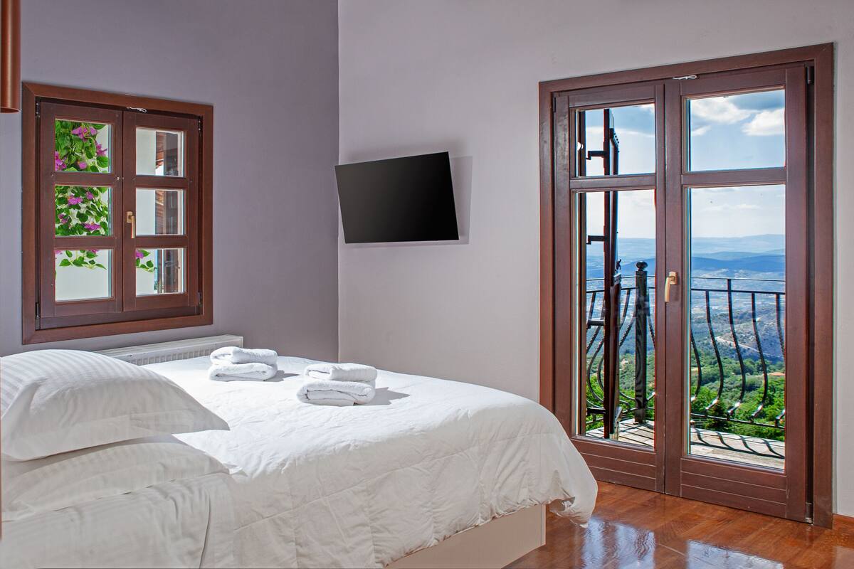 A bedroom with a large bed, a TV mounted on the wall, and a balcony with a view of the landscape.