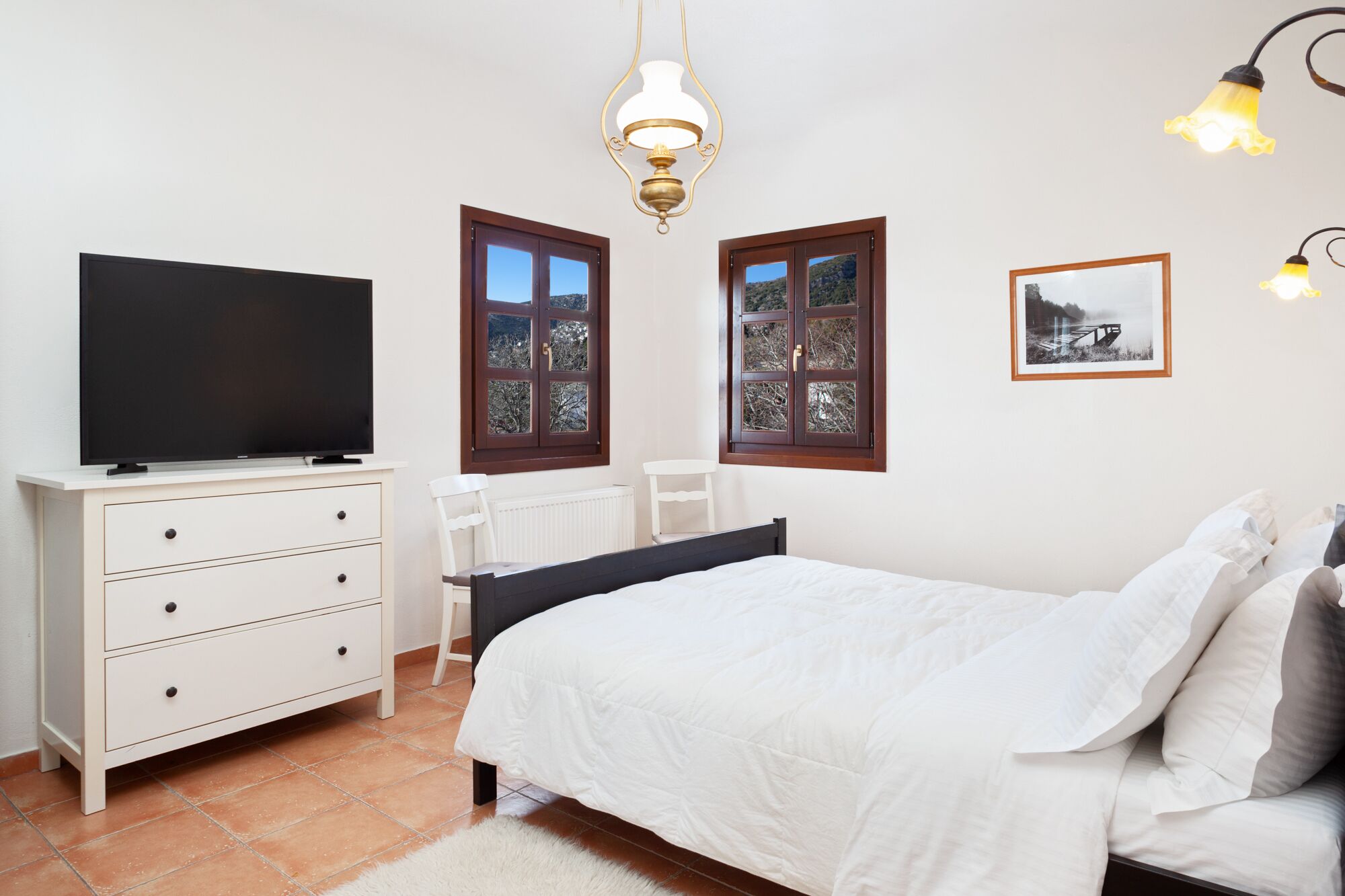 A bedroom with a bed, a TV on a dresser, two windows, and a picture on the wall.