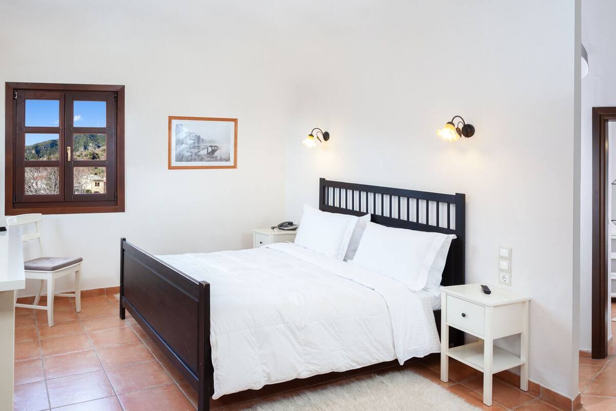 A bedroom with a bed, a desk, a chair, a TV, a window, a bedside table, a nightstand, a lamp, and a mirror.