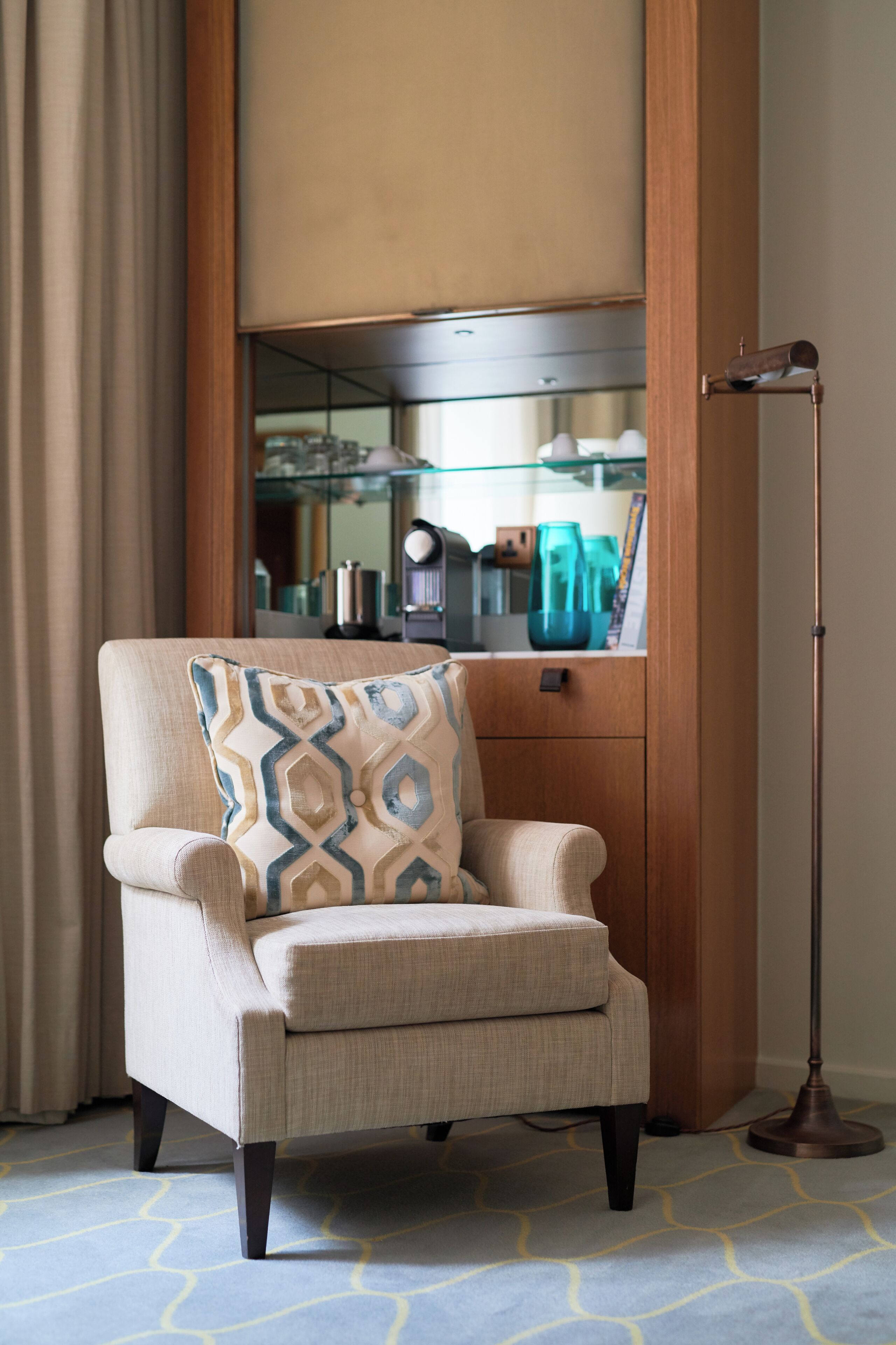 A corner room with a beige armchair, a patterned pillow, a floor lamp, a mirror, and a glass cabinet with decorative items.