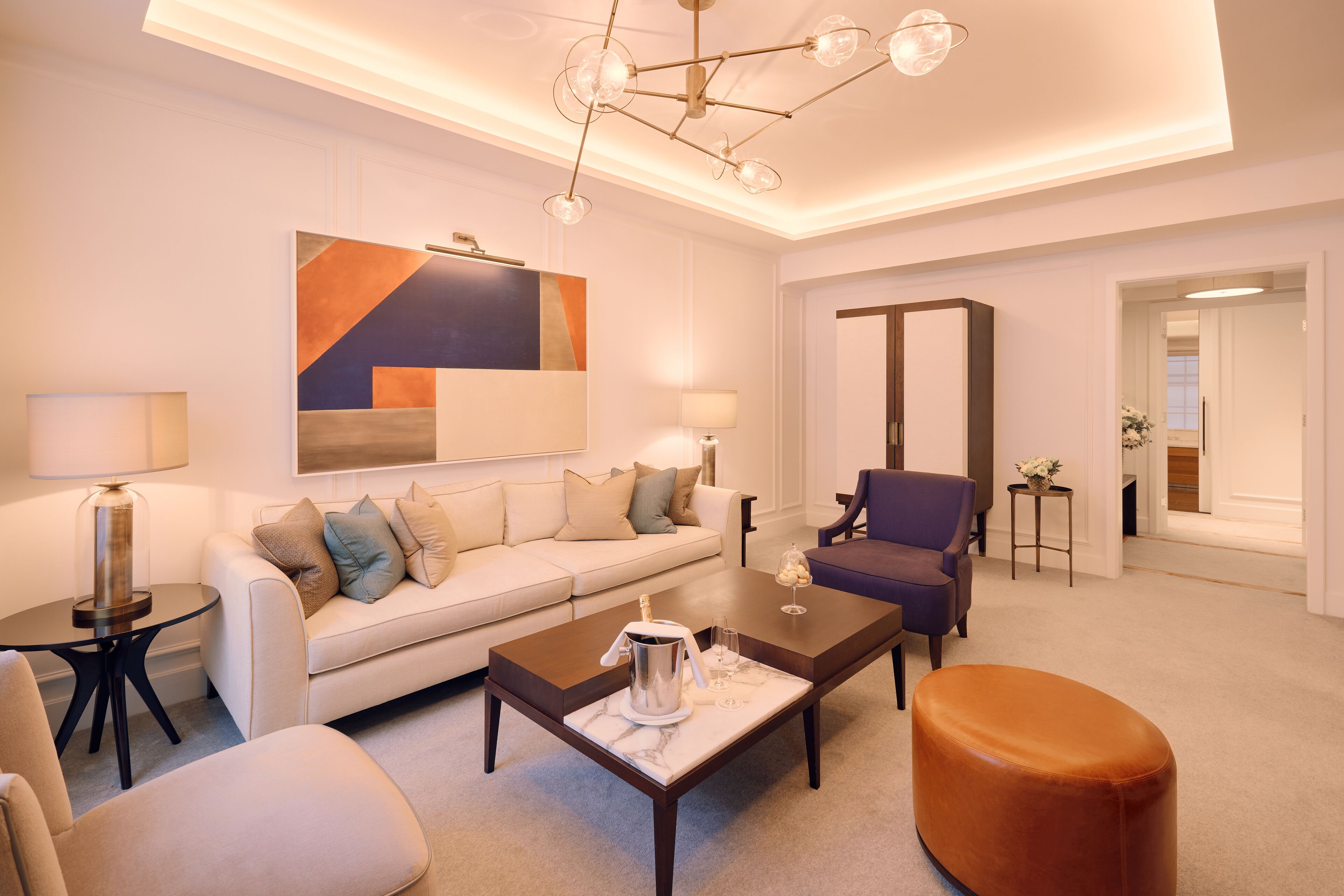 A modern living room with a sofa, coffee table, and armchairs. There is a painting on the wall and a chandelier above the seating area.