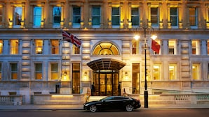 A grand building with a large entrance, a flag, and a car parked in front. - Corinthia London (London)