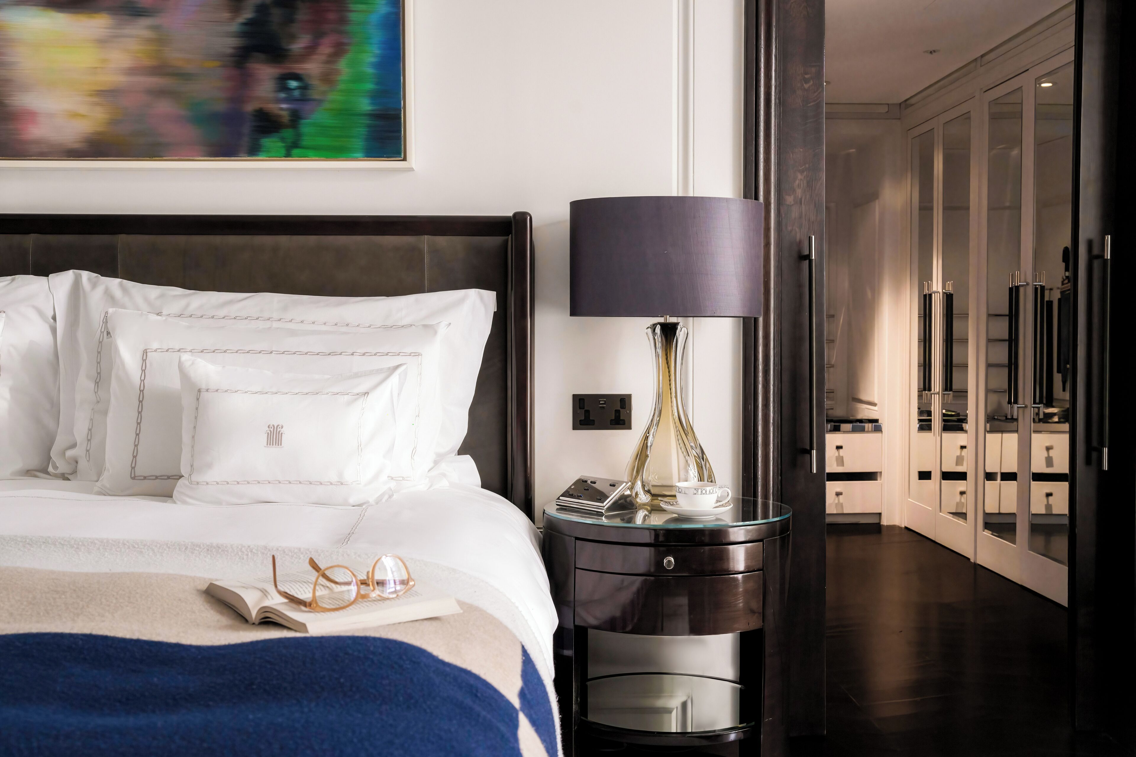 Una cama bien hecha con ropa blanca, una mesita de noche con lámpara y un aparador de vidrio con cajones.