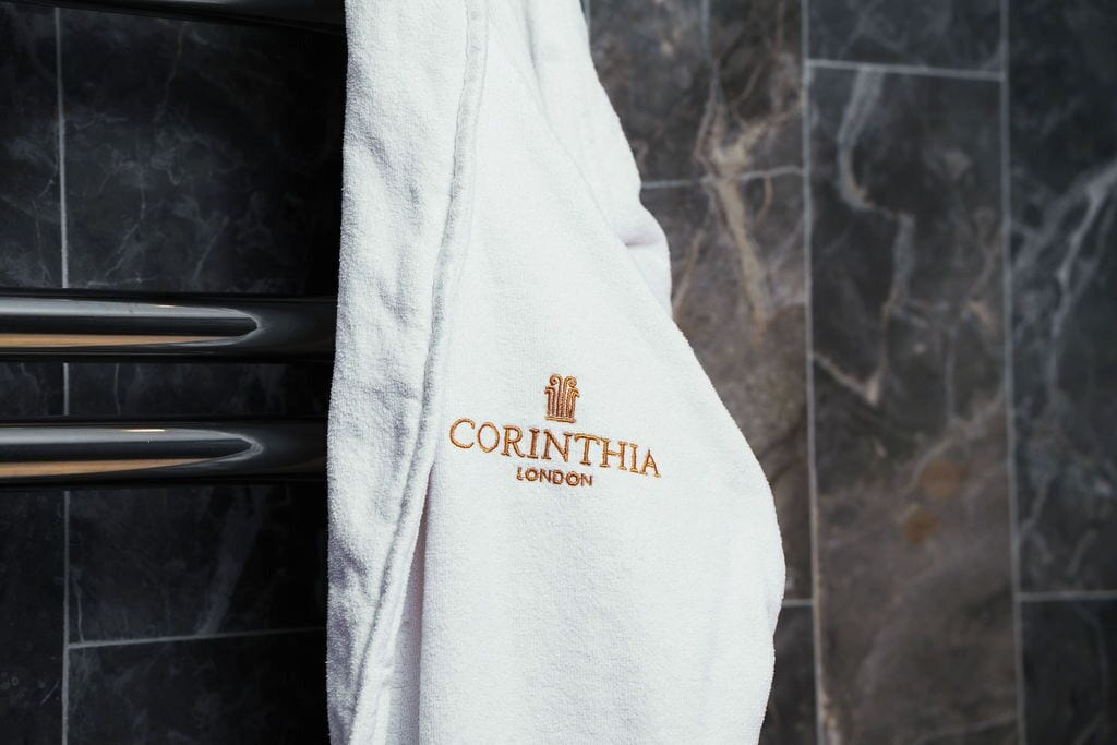 A white towel with the text "CORINTHIA LONDON" hanging on a black tiled wall.