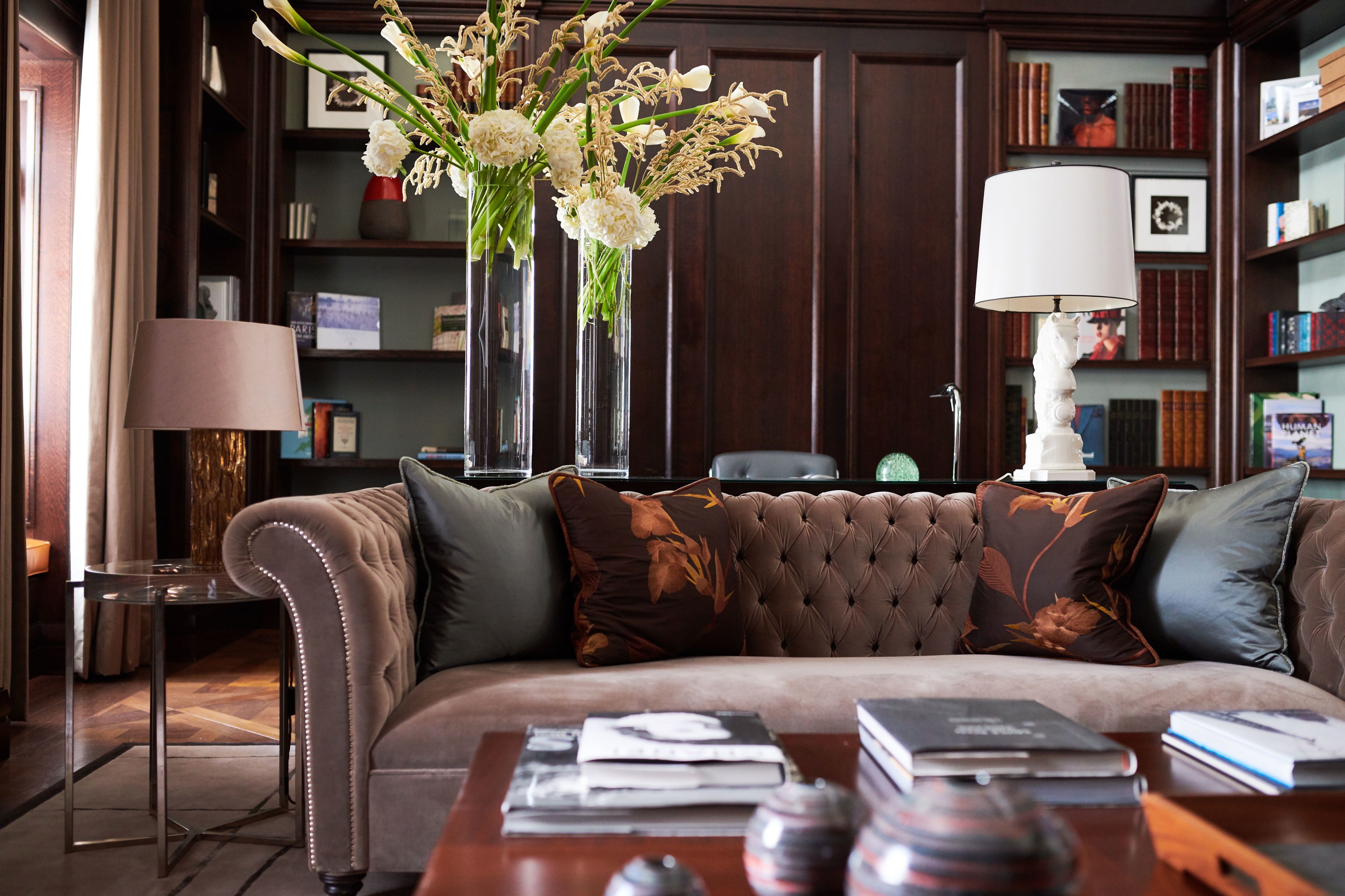 A plush brown sofa with decorative pillows, a side table with a lamp, and a vase with white flowers.