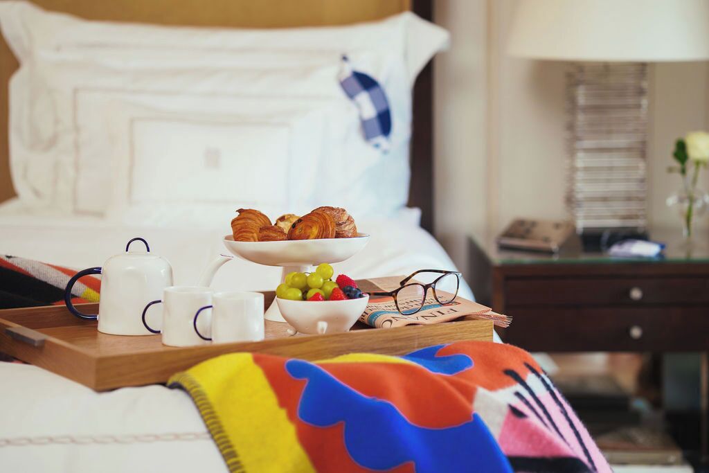A neatly made bed with a tray of breakfast items including croissants, a bowl of fruit, and white mugs.
