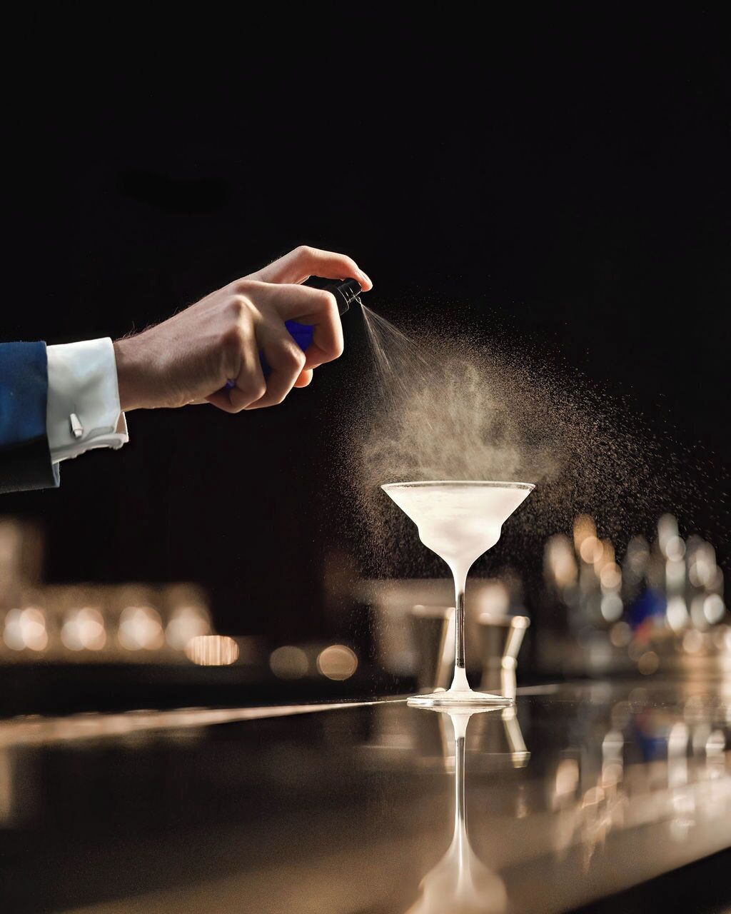 A hand sprinkling a powdery substance over a martini glass on a bar counter.