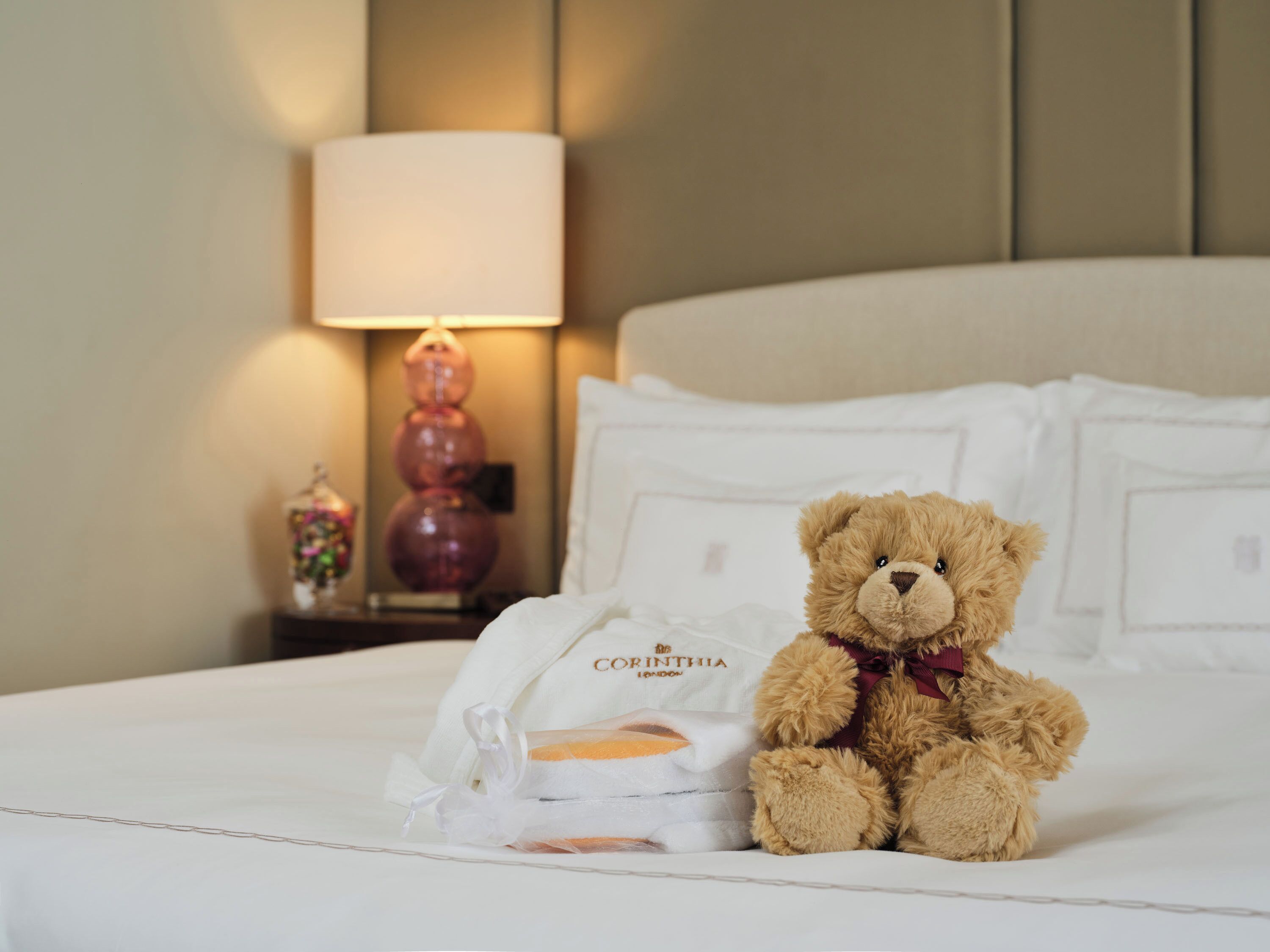 A hotel room with a bed, a teddy bear, a lamp, and a nightstand with a decorative item.