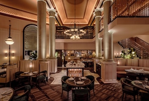 A spacious hotel lobby with a central dining area, multiple seating options, and a staircase leading to an upper level.