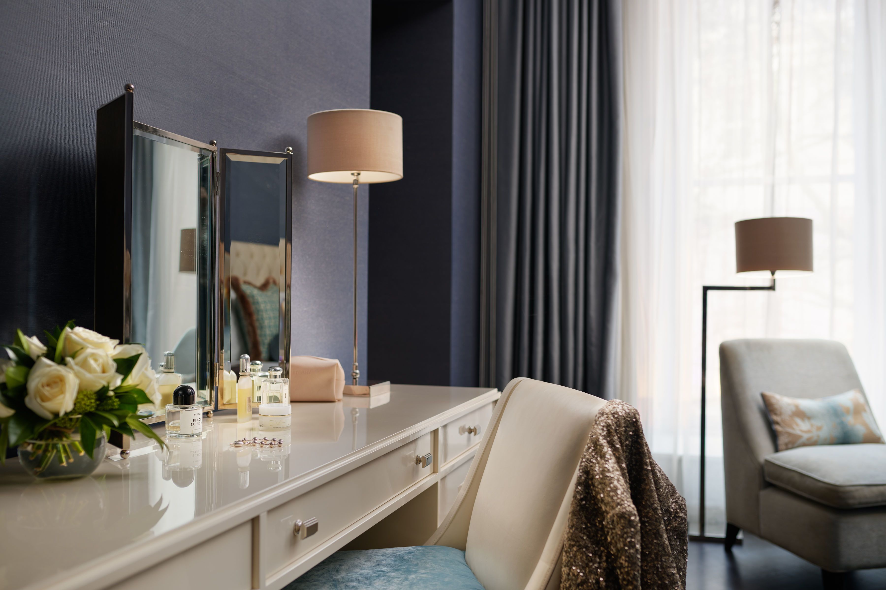 Een hotelkamer met een kaptafel, spiegel, lamp, stoel en raam met gordijnen.