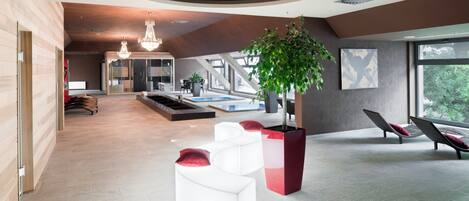 A spacious indoor area with a modern design, featuring a large open space, a seating area with white and red cushions, a small indoor garden, and a view through large windows.