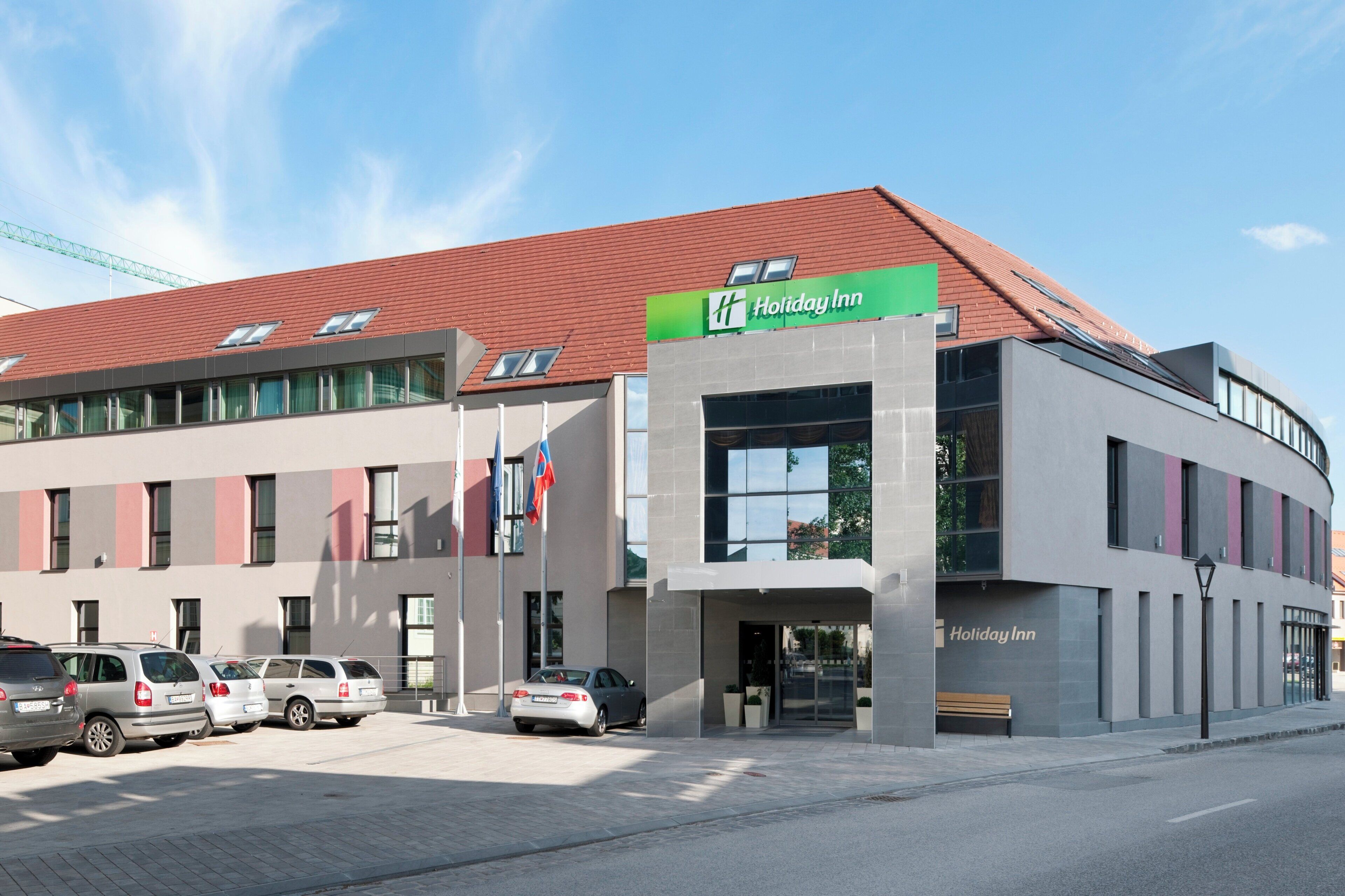 Un hotel Holiday Inn con diseño moderno, que cuenta con un techo rojo, ventanas amplias y un estacionamiento con varios autos.