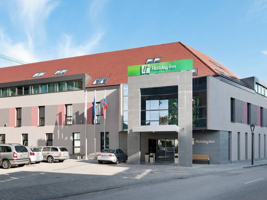 Un hotel Holiday Inn con diseño moderno, que cuenta con un techo rojo, ventanas amplias y un estacionamiento con varios autos.