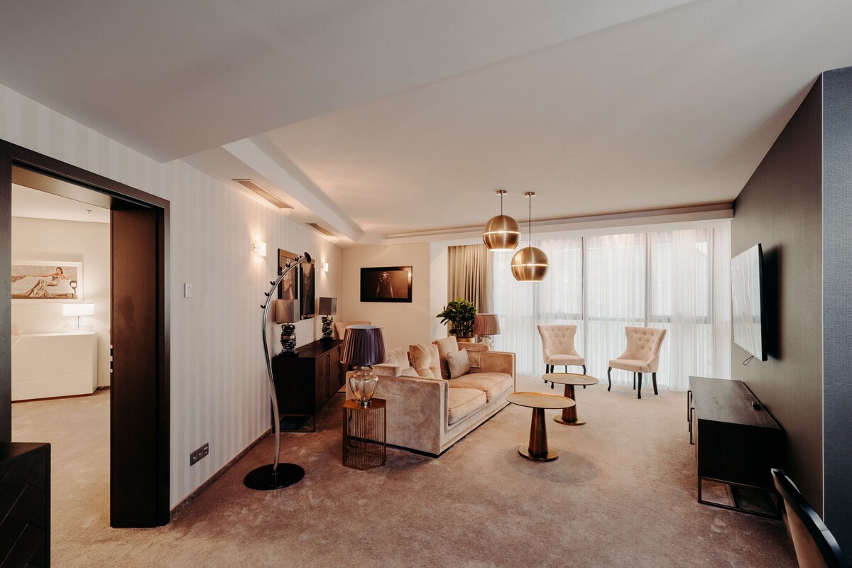 Een moderne woonkamer met een bank, stoelen, een salontafel en een televisie. Een deur leidt naar een andere kamer, en een lamp is zichtbaar.