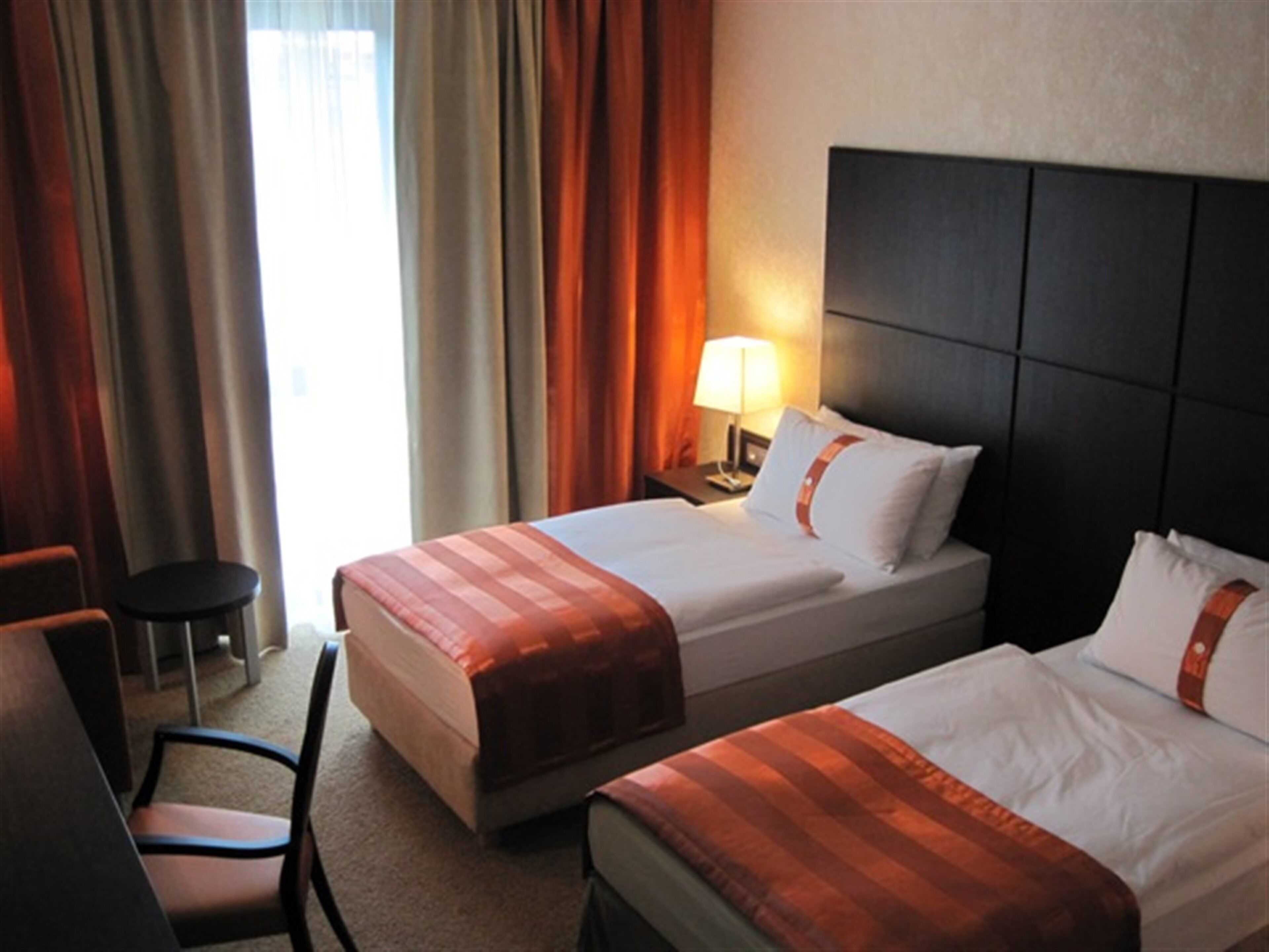 Habitación de hotel con dos camas, una silla, una mesita de noche con lámpara y una ventana con cortinas.