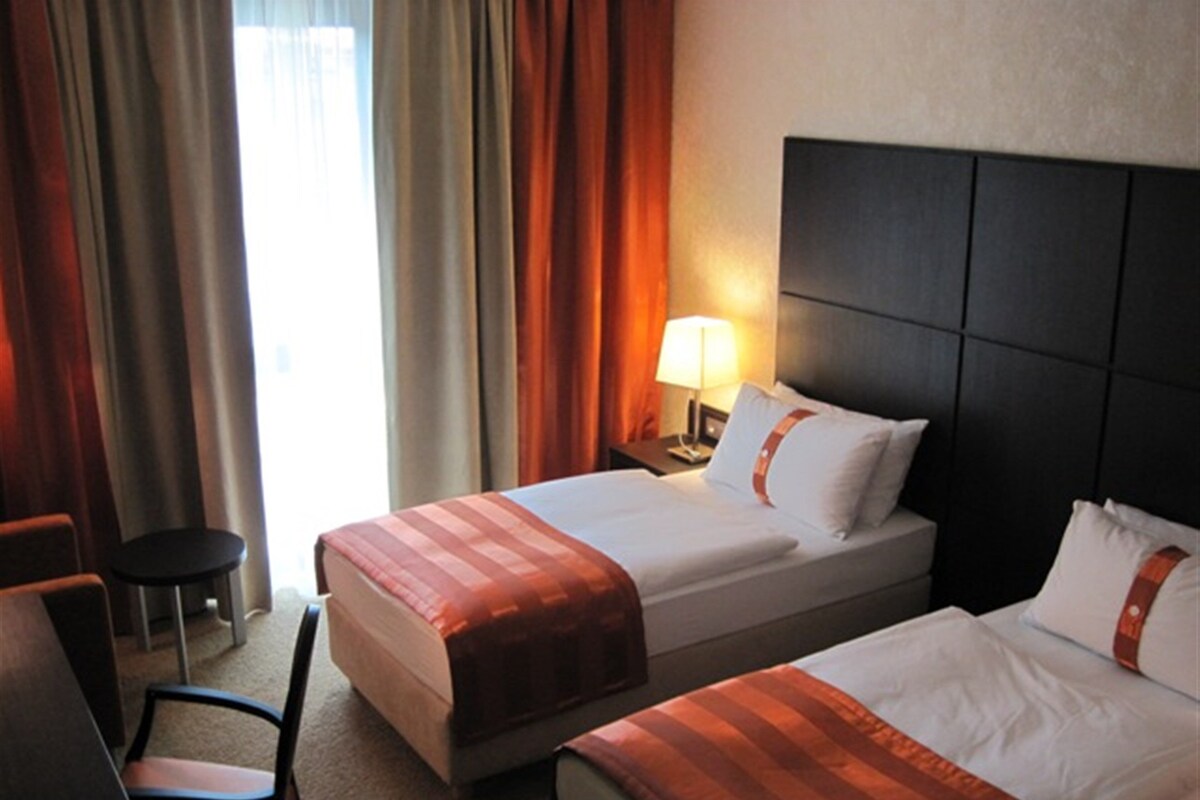Een hotelkamer met twee bedden, een stoel, een nachtkastje met lamp en een raam met gordijnen.
