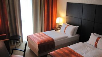 Habitación de hotel con dos camas, una silla, una mesita de noche con lámpara y una ventana con cortinas.