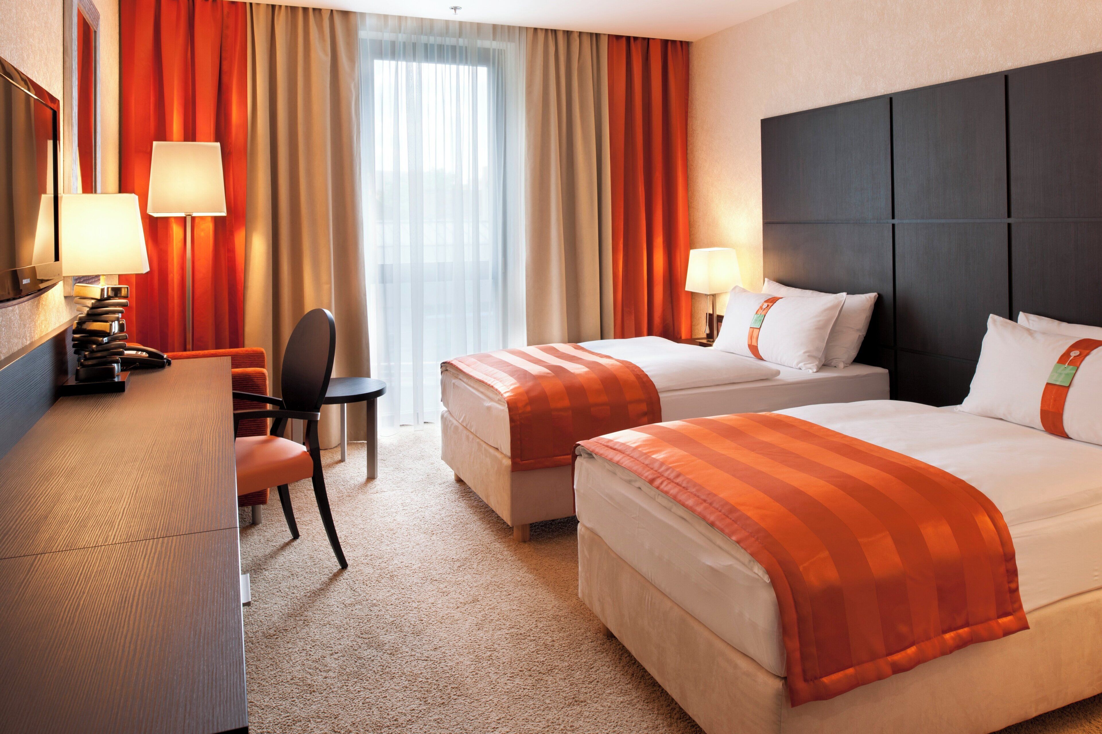 A hotel room with two beds, a desk, a chair, and a lamp.