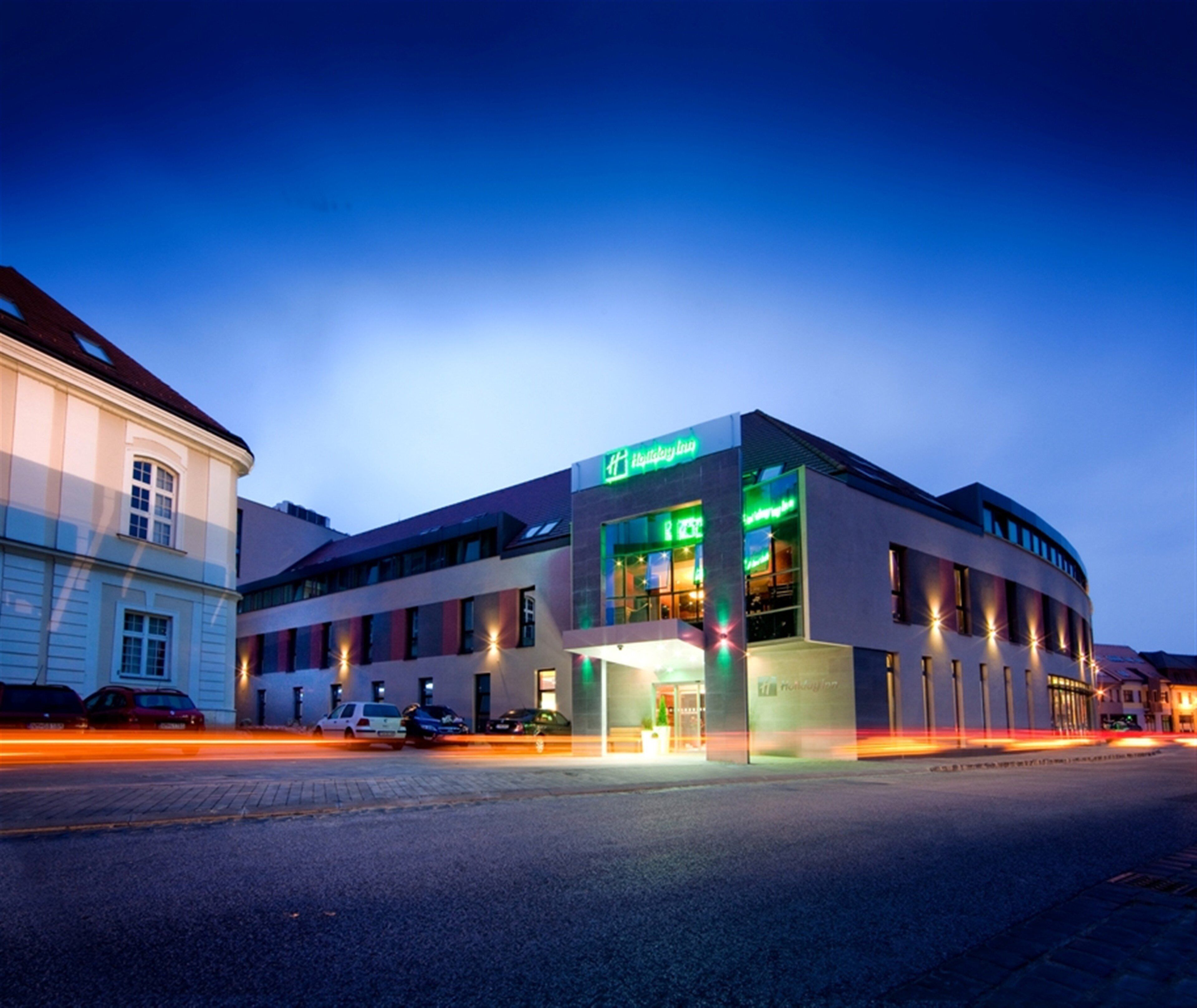 A Holiday Inn hotel with a modern design, featuring a prominent green sign, multiple windows, and a parking lot in the foreground.