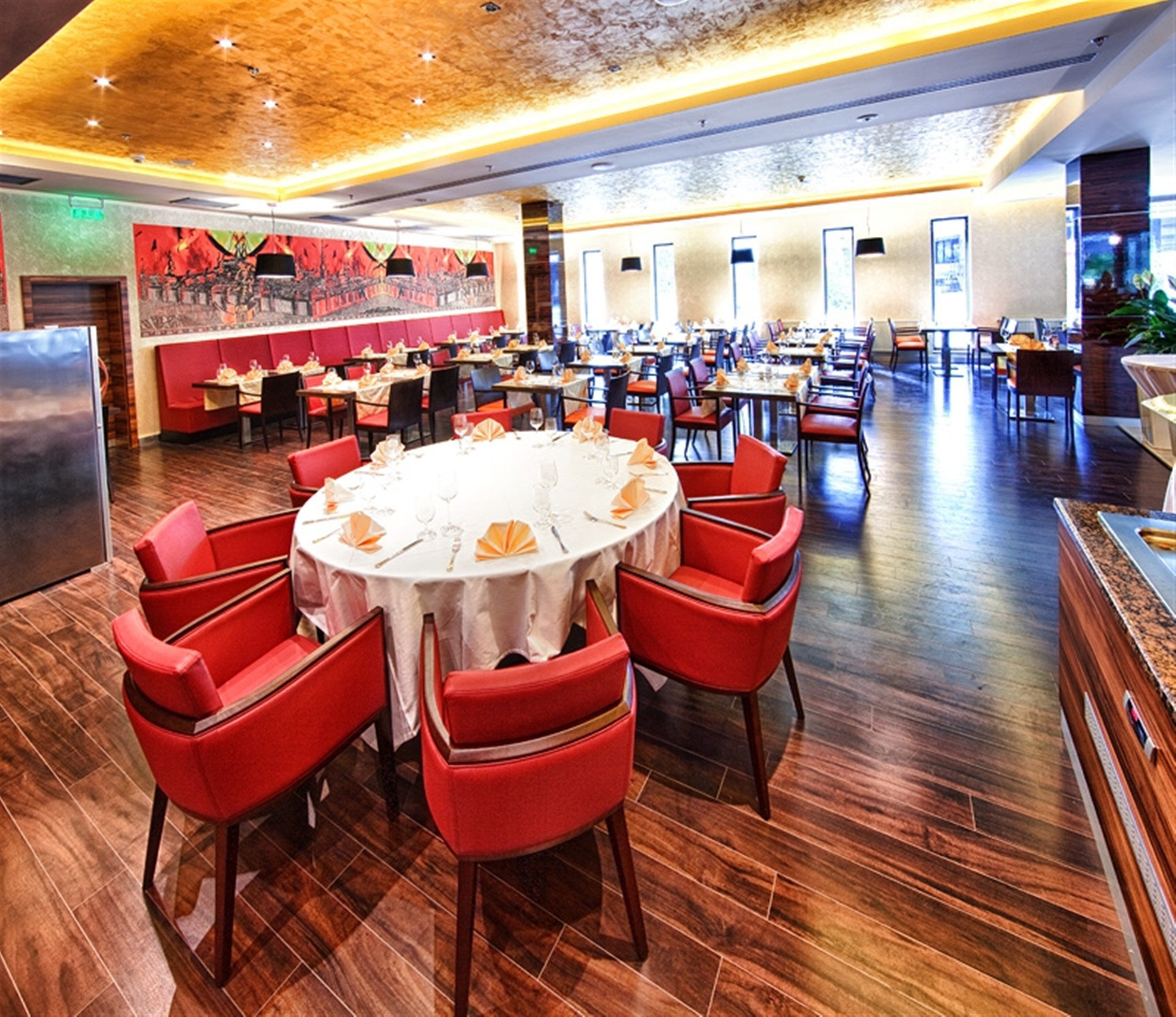 A spacious dining area with round tables and red chairs, a large mural on the wall, and a modern ceiling design.