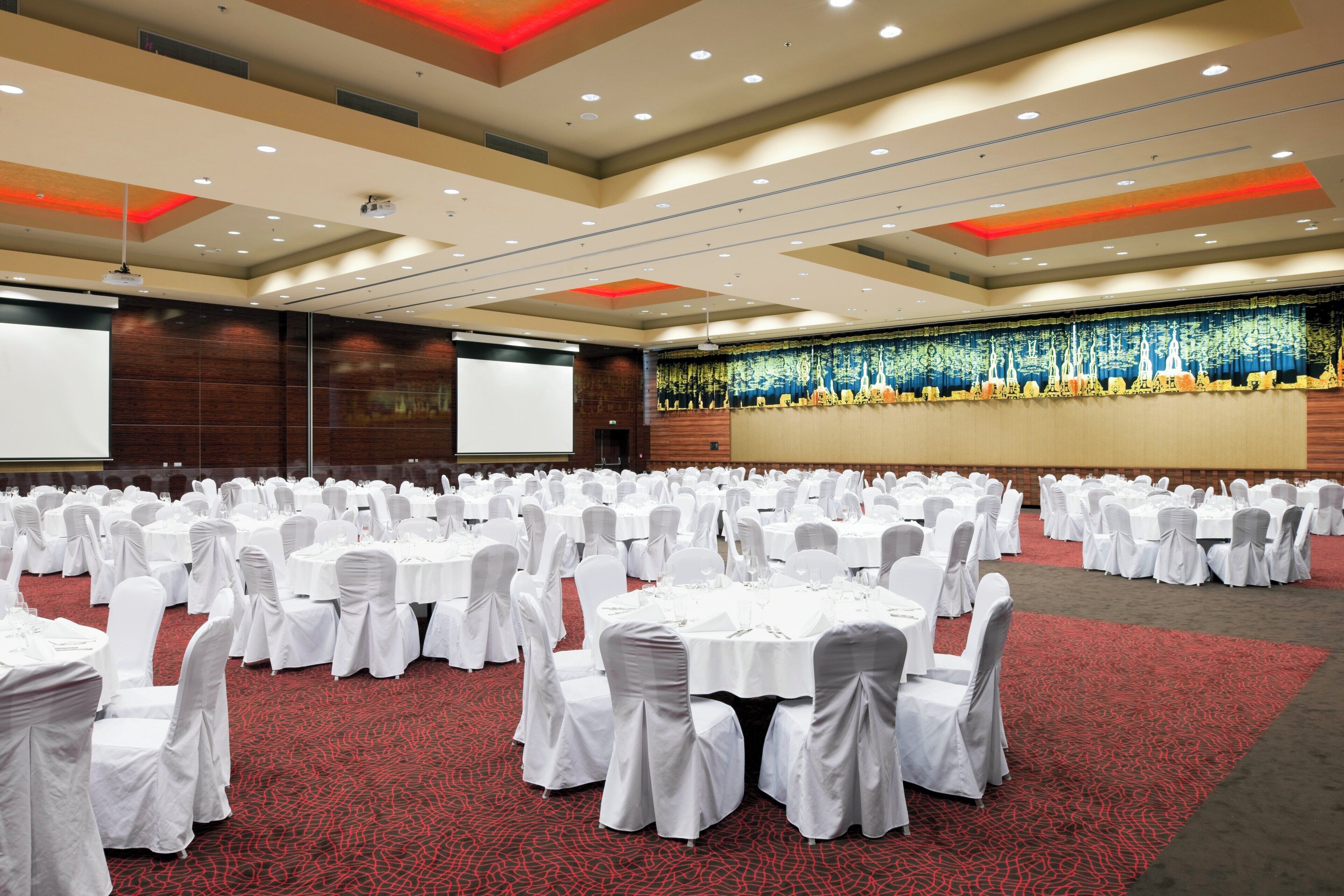 A large conference room with round tables and chairs, a projector screen, and a stage with a backdrop.