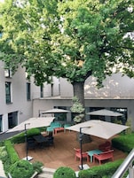 Un área de patio al aire libre con piso de madera, varias mesas y sillas, y sombrillas que dan sombra.