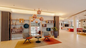 A modern lobby with a seating area featuring two armchairs and a small table, a wooden reception desk, and a large potted plant.