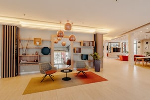 Un vestíbulo moderno con zona de estar que incluye dos sillones y una mesita, un mostrador de recepción de madera y una planta grande en maceta.
