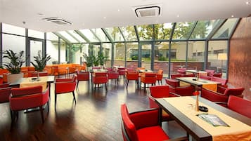 A modern restaurant with red chairs and tables, large windows, and indoor plants.