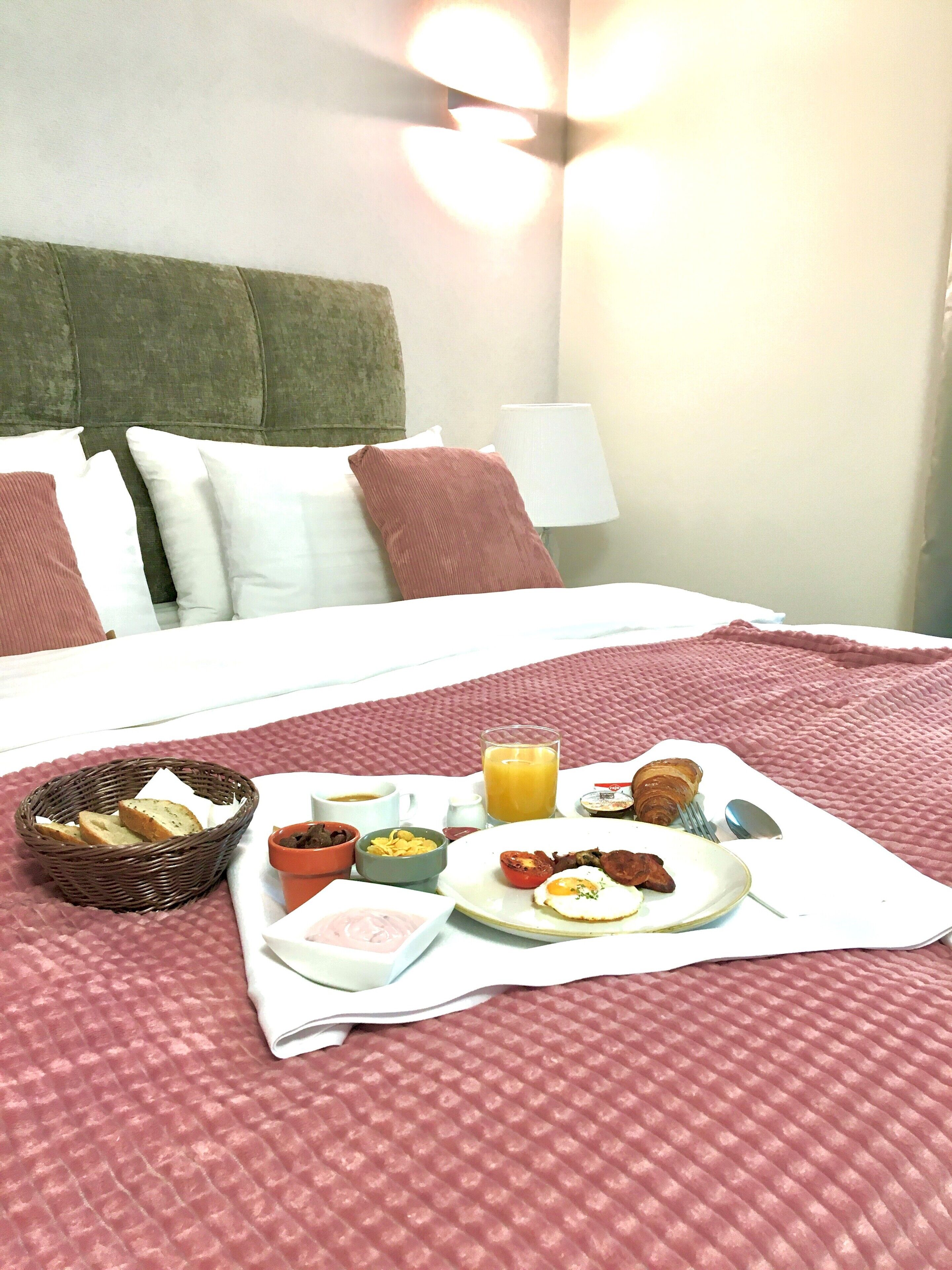 Un lit bien fait, recouvert d’une couverture rouge, une table de chevet avec une lampe, et un plateau de petit-déjeuner avec des toasts, des œufs et du jus d’orange.