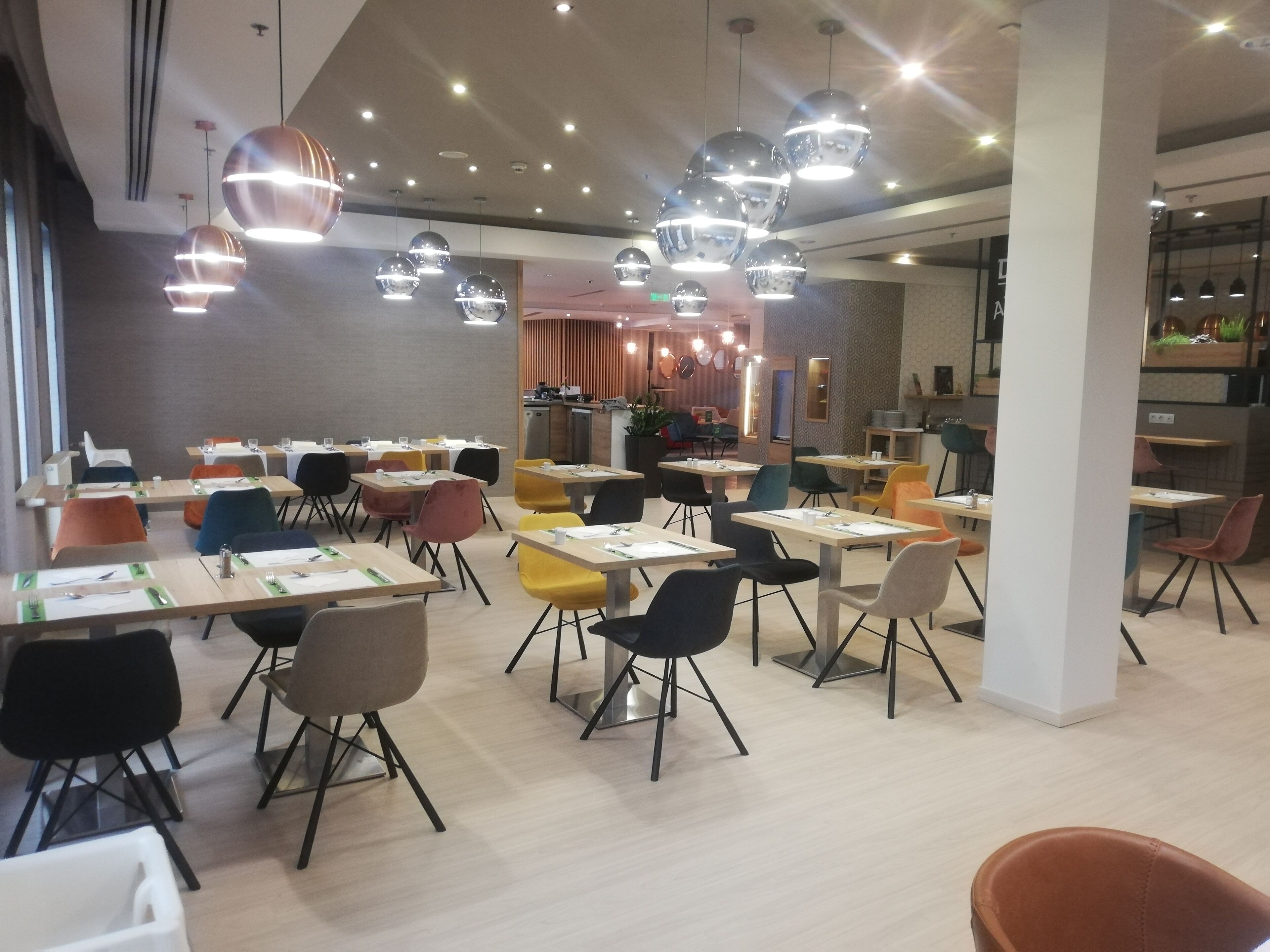 A modern dining area with multiple tables and chairs, overhead lighting, and a kitchen area in the background.
